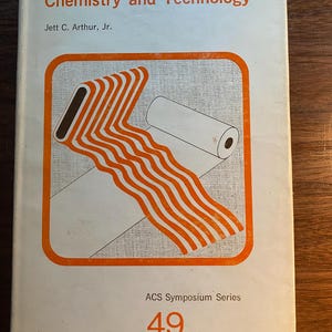 May include: A white book cover with orange text that reads "Textile and Paper Chemistry and Technology" by Jett C. Arthur, Jr. The cover also features an orange and white illustration of a roll of paper and a ribbon.