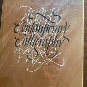 May include: A book cover with a gold-toned background and the title "Modern Scribes and Lettering Artists II." The cover features the words "Alphabet, Contemporary, Calligraphy, Skills" in a mix of black and white stylized calligraphy.