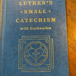 May include: A blue hardcover book titled Luther's Small Catechism with Explanation in gold lettering. The cover features a gold embossed Luther Rose emblem consisting of a heart, a cross, and a circular floral border within a square frame.