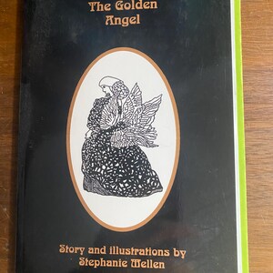 Puede incluir: Un libro de tapa dura titulado "The Golden Angel" con una cubierta negra y una ilustración de un ángel en un marco ovalado. El lomo del libro es verde y blanco. El texto de la portada dice "Story and illustrations by Stephanie Wellen."
