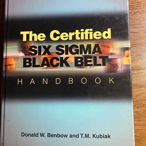 May include: A book titled "The Certified Six Sigma Black Belt Handbook" with a gradient blue and black cover. The title is in yellow and black font. The authors' names, Donald W. Benbow and T.M. Kubiak, are printed at the bottom.