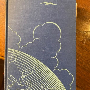 Op de afbeelding: Een blauw hardback boek met een groene en witte illustratie van een wereldbol en wolken op de omslag. De wereldbol wordt omringd door een witte vogel die in de lucht vliegt.