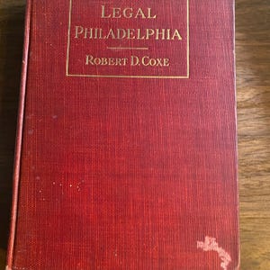 May include: A vintage book titled "Legal Philadelphia" by Robert D. Coxe. The book has a textured, deep red cover with gold lettering. The title and author's name are enclosed in a gold rectangular frame.