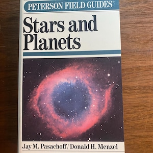 May include: A softcover book titled Peterson Field Guides Stars and Planets by Jay M. Pasachoff and Donald H. Menzel. The white cover features a large, circular, pink and blue nebula against a black starfield, with dark teal horizontal bars.