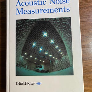 Könnte beinhalten: Ein weißes Buch mit dem Titel "Acoustic Noise Measurements" in blauer Schrift. Das Cover zeigt ein Foto eines schalltoten Raums mit einem LKW darin. Das Buch wurde von Brüel & Kjær herausgegeben.