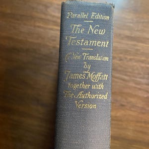 Puede incluir: Primer plano del lomo de un libro con letras doradas sobre una cubierta gris. El texto dice "The New Testament" e incluye "Parallel Edition", "New Translation" y "Authorized Version".