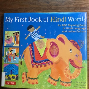 Puede incluir: Un libro infantil titulado "My First Book of Hindi Words" con una cubierta azul. La portada presenta un elefante naranja, un niño e ilustraciones de la cultura india. El libro es de Rina Singh e ilustrado por Farida Zaman.