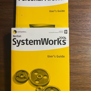 May include: Two yellow Norton software manuals, "Personal Firewall 2004" and "SystemWorks 2004", are stacked on a wooden surface. The manuals have white and black text, with the "User's Guide" title visible.