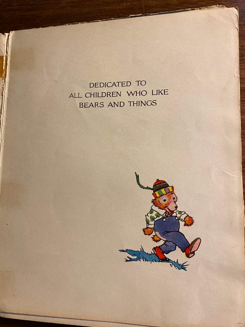 K&ouml;nnte beinhalten: Eine Widmungsseite aus einem Kinderbuch mit dem Text "Gewidmet allen Kindern, die B&auml;ren und Dinge m&ouml;gen". Die Seite zeigt eine Comic-Illustration eines B&auml;ren, der einen gr&uuml;n-gelb-roten Hut, eine blaue Latzhose und ein wei&szlig;es Hemd mit gr&uuml;nen Punkten tr&auml;gt. Der B&auml;r geht auf einem blau-gr&uuml;nen Weg.