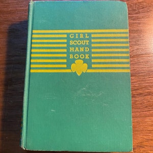 May include: A green book with the title "Girl Scout Handbook" in yellow letters on the front cover. The book has a yellow and green striped design and a Girl Scout emblem.