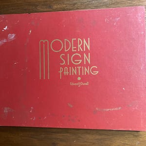 May include: A red book with the title "Modern Sign Painting" in gold lettering. The cover features a vintage design, with the author's name, Edward J. Duvall, printed below the title. The book is on a wooden surface.