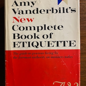May include: A red hardcover book titled "Amy Vanderbilt's New Complete Book of Etiquette" with the tagline "The guide to gracious living by the foremost authority on manners today". The book has a white spine with a blue and red stripe. The bottom of the cover has the text "Over 1,300,000 COPIES IN USE" and the initials "AV" in cursive.