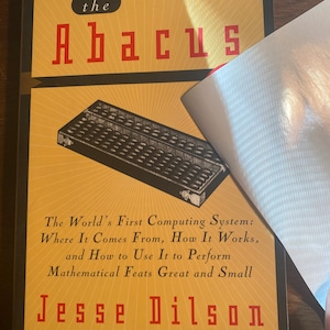 Puede incluir: Un libro titulado "The Abacus: The World's First Computing System: Where It Comes From, How It Works, and How to Use It to Perform Mathematical Feats Great and Small" de Jesse Dilson. La portada presenta un ábaco en blanco y negro.