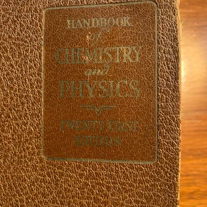 May include: Close-up of a vintage book titled "Handbook of Chemistry and Physics, Twenty First Edition." The cover is a textured brown with a rectangular label in a lighter shade of brown. The text is in a green font.