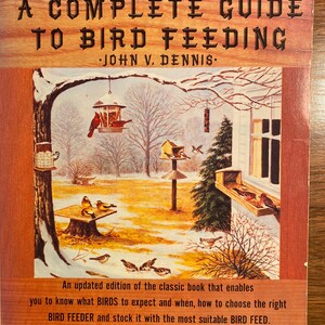 Può includere: Una copertina di libro vintage con una scena invernale con uccelli a un mangiatoia. Il titolo del libro è "A Complete Guide to Bird Feeding" di John V. Dennis.
