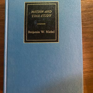 May include: A blue hardcover book titled Motion and Time Study by Benjamin W. Niebel. The front cover features a central black rectangular label with gold lettering and a thin gold border, set against a solid blue textured background.