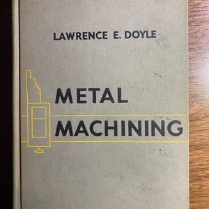 May include: A hardcover book titled "METAL MACHINING" by Lawrence E. Doyle. The book has a light gray cover with black text and a yellow illustration of a machine. The book is lying on a wooden surface.
