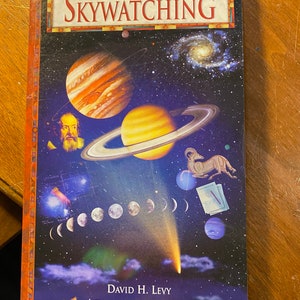 Op de afbeelding: De omslag van het boek Skywatching van The Nature Company Guides toont een kleurrijke illustratie van planeten, sterren en een komeet tegen een donkerblauwe achtergrond. De titel "Skywatching" staat in grote witte letters. De auteur is David H. Levy met een inleiding van Robert Burnham.