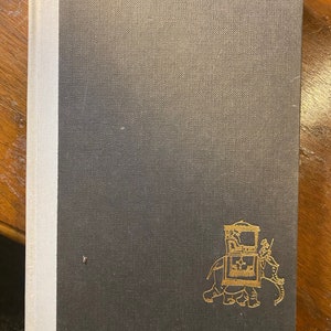 May include: A black hardcover book with a gold-colored illustration of an elephant carrying a palanquin on its back. The book has a white spine.