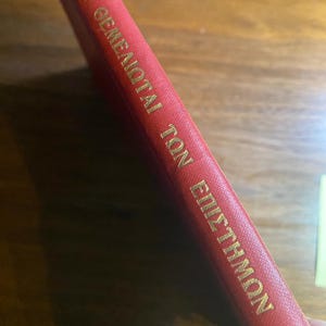 Könnte beinhalten: Ein rotes Buch mit goldener Schrift. Der Text auf dem Buchrücken lautet "ΟΙ ΘΕΜΕΛΙΩΤΑΙ ΤΩΝ ΕΠΙΣΤΗΜΩΝ". Das Buch liegt auf einer Holzoberfläche.