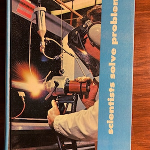 Op de afbeelding: Een boekomslag met een blauwe achtergrond en de tekst "scientists solve problems" in wit. De omslag toont een foto van een persoon die een lasmasker draagt en een lasbrander vasthoudt. De tekst "SINGER SCIENCE SERIES" staat onderaan de omslag.