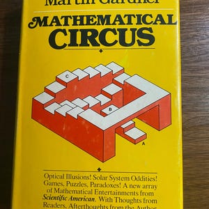 Może przedstawiać: Książka w stylu vintage zatytułowana "Mathematical Circus" autorstwa Martina Gardnera. Okładka jest żółta z czerwonymi i białymi geometrycznymi kształtami i tekstem. Książka obiecuje iluzje optyczne, łamigłówki i rozrywkę matematyczną.