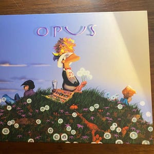 Op de afbeelding: Albumhoes met het woord "OPUS" in paars. De illustratie toont een cartoon eend met een hoed en een "25 YEARS" sjerp, zittend op een grasheuvel met andere cartoonfiguren en paardenbloemen.
