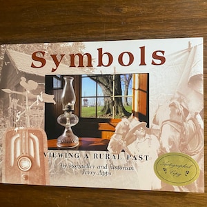 May include: A book titled Symbols: Viewing a Rural Past by Jerry Apps. The cover features a sepia-toned collage of a weather vane, a radio, a horse, and a person, with a central color photograph of a glass oil lamp in front of a window.