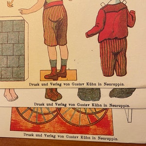 May include: A vintage paper doll set featuring a boy wearing a red and white striped outfit. The paper doll is standing on a platform with the text "Druck und Verlag von Gustav K&uuml;hn in Neuruppin." printed below.