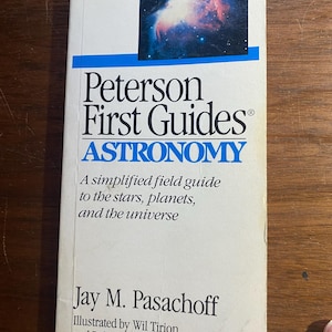 Puede incluir: Un libro de bolsillo titulado "Peterson First Guides: Astronomy". La portada presenta una imagen azul y blanca de una nebulosa. Es una guía de campo simplificada de estrellas, planetas y el universo, escrita por Jay M. Pasachoff.