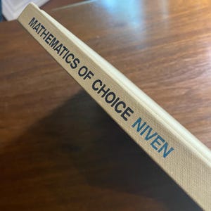 Op de afbeelding: Een hardcover boek getiteld "Mathematics of Choice Niven" met een beige omslag. De titel en de naam van de auteur zijn in donkerblauw gedrukt. Het boek ligt op een donkerhouten oppervlak.