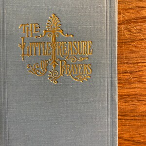 May include: A light blue book titled "The Little Treasure of Prayers" with gold lettering and decorative flourishes. The book has a textured cover and a rectangular shape.
