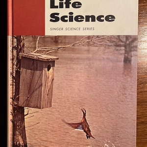 May include: A vintage book titled "Basic Life Science" from the Singer Science Series. The cover features a brown wooden birdhouse on a tree branch over a body of water with a duck taking flight.