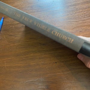 May include: A book spine with the title "THE TRUE VISIBLE CHURCH" in gold lettering. The book has a grey-green cover and a black spine. The book is resting on a wooden surface.