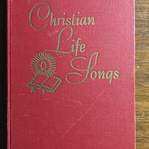 Puede incluir: Un libro de tapa dura rojo con el título "Christian Life Songs" en letras doradas. La portada presenta una ilustración dorada de un libro con una vela y un sol.