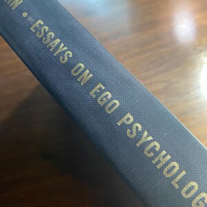May include: Close-up of a book spine with the title "ESSAYS ON EGO PSYCHOLOGY" in gold lettering. The book has a dark blue cover. The book is angled, revealing the title and author's name.