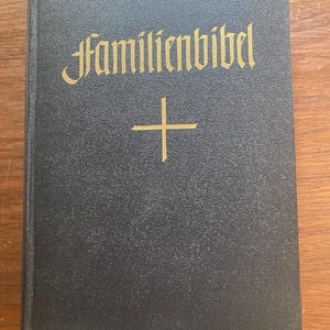 以下が含まれることがあります： 金色の「Familienbibel」の文字と金の十字架が入った黒いハードカバーの本。本はテクスチャ加工された表紙で、木製の表面に置かれています。