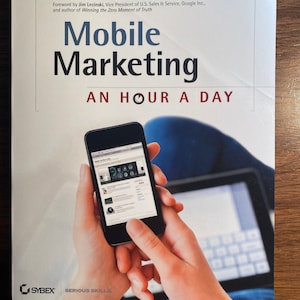 May include: A book titled "Mobile Marketing: An Hour a Day" by Rachel Pasqua and Noah Elkin. The cover features a person holding a smartphone with a website open on the screen. The book is published by Sybex and is part of their "Serious Skills" series.