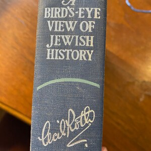 Op de afbeelding: Een blauw boek met de titel "A Bird's-Eye View of Jewish History" en een handtekening die "Cecil Roth" in zilveren letters luidt.