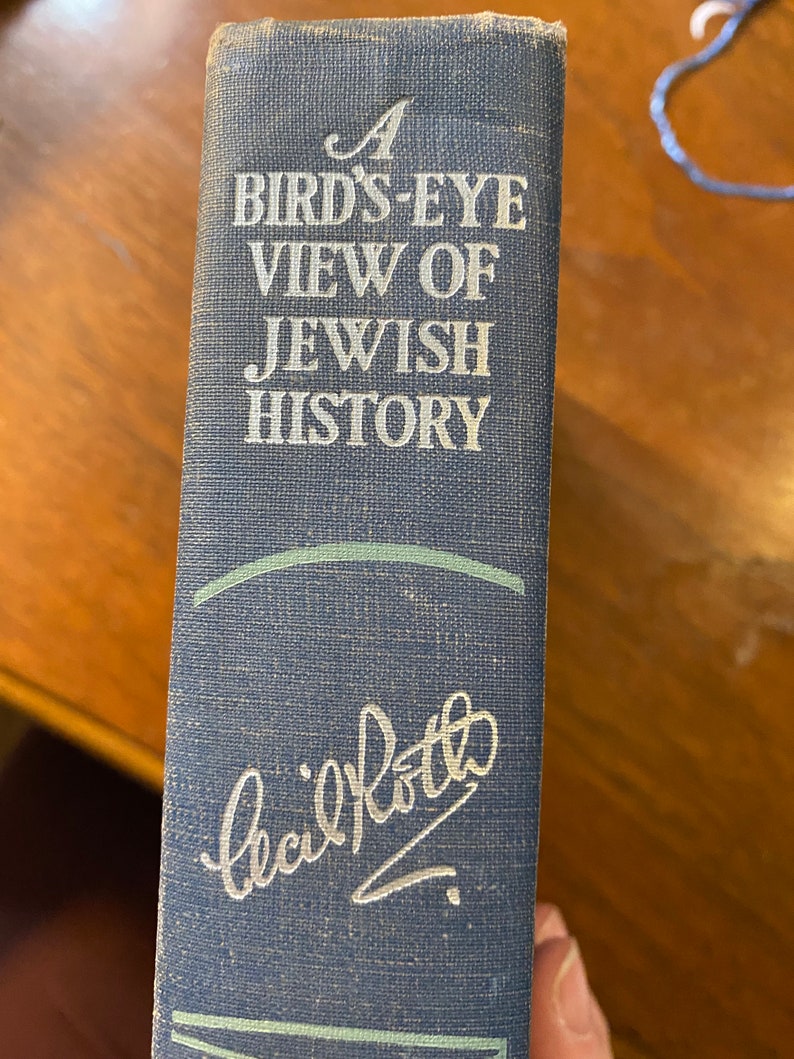 Op de afbeelding: Een blauw boek met de titel "A Bird's-Eye View of Jewish History" en een handtekening die "Cecil Roth" in zilveren letters luidt.