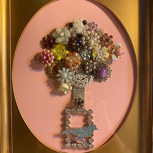 May include: A framed artwork featuring a colorful bouquet of vintage jewelry pieces arranged to resemble a tree, with a bird-shaped brooch at the base.
