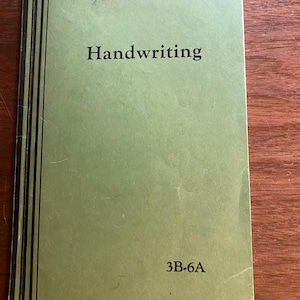 May include: A green book with black stripes on the spine. The book is titled "Handwriting" and has the text "3B-6A" on the bottom right corner. The book has a handwritten name on the top right corner.