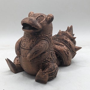 May include: A 3D printed bronze-coloured figurine of a Pokemon character. The character is sitting with its legs crossed and has a large, spiky tail.