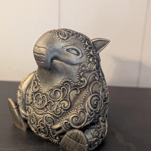 Puede incluir: Figura de oveja dorada con intrincados patrones florales arremolinados. La oveja está sentada, con las patas recogidas. La escultura tiene una superficie texturizada y pequeñas orejas puntiagudas. Acabado metálico.