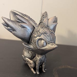 May include: A silver-colored figurine of a stylized fox-like creature. The figurine has large, pointed ears and intricate, ornate detailing around the head, chest, and legs. The creature is in a seated position.