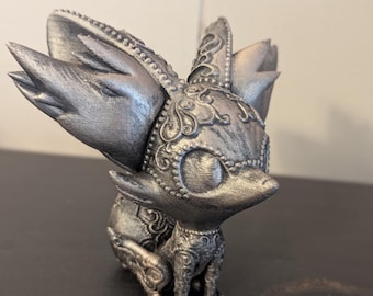 May include: A silver-colored figurine of a stylized fox-like creature. The figurine has large, pointed ears and intricate, ornate detailing around the head, chest, and legs. The creature is in a seated position.