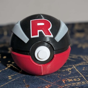 Puede incluir: Réplica impresa en 3D de una Poké Ball del Team Rocket. La bola es principalmente negra con una base roja y un botón blanco. La letra "R" se muestra en rojo en la mitad superior negra, con detalles grises.