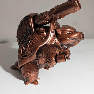 May include: A bronze-colored sculpture of a turtle with a cannon mounted on its back. The turtle has detailed armor and a smiling face. The cannon is also intricately designed, with a textured surface and a flared muzzle. The sculpture is on a white surface.