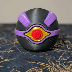 Puede incluir: Objeto esférico negro con detalles morados y un diseño central en forma de ojo en oro, rojo y amarillo. El objeto tiene una superficie texturizada y parece estar impreso en 3D. El diseño recuerda a un accesorio de fantasía o ciencia ficción.