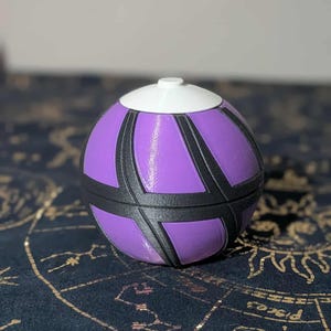 Puede incluir: Un recipiente esférico con tapa blanca y cuerpo morado. El cuerpo está decorado con líneas negras y curvas. El recipiente está sobre una superficie azul oscuro con símbolos astrológicos dorados.